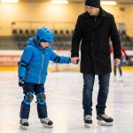 Un père tient la main de son enfant protégé par un casque et des genouillères lors d'une séance de patinage sur glace.